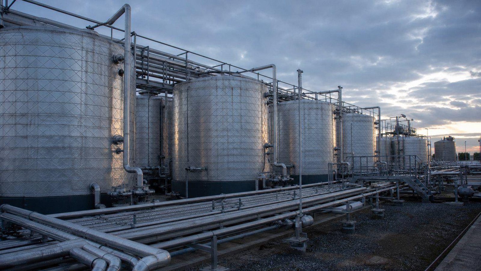 Row of stainless steel chemical storage tanks with pipe racks under a cloudy sky