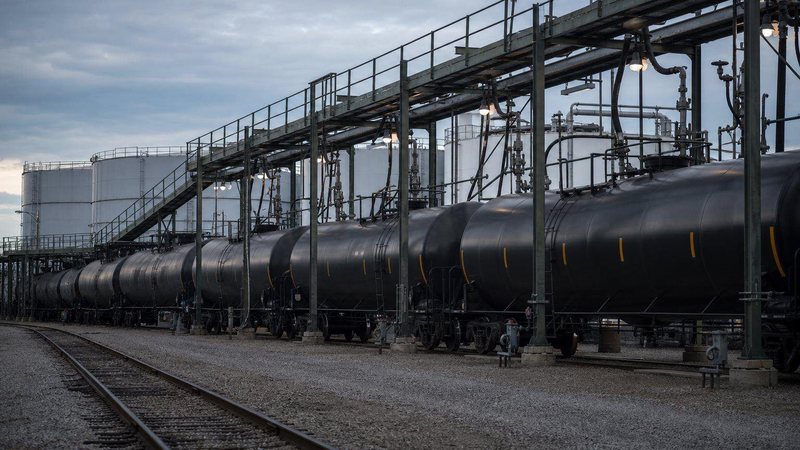 Unit train of black rail tank cars at a loading rack beside storage tanks