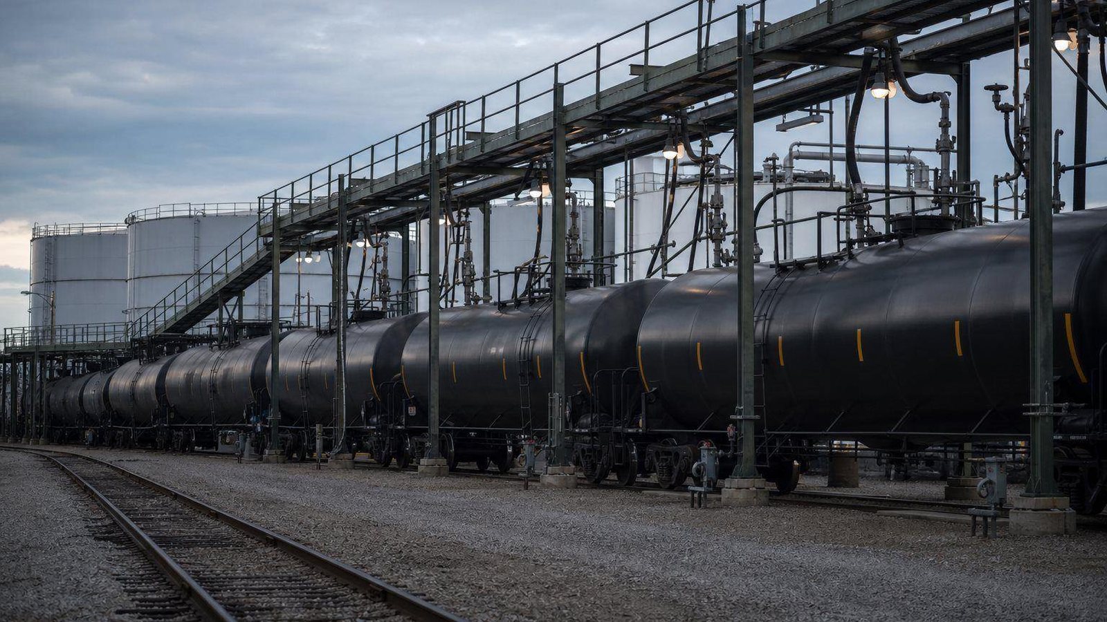 Unit train of black rail tank cars at a loading rack beside storage tanks